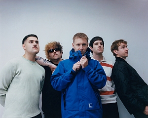 Five men stand against a plain white wall; one wears a blue jacket and zips it up, while others stand casually, one with sunglasses and another in a striped shirt.