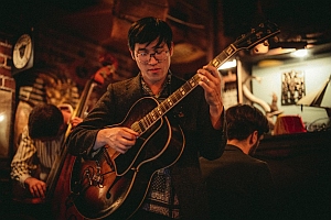 A man plays an electric guitar in a dimly lit room with two other musicians in the background, one playing a double bass and one at a piano.