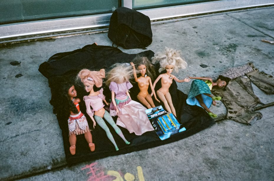 Six dolls, some clothed and some unclothed, are laid out on a black cloth on a concrete sidewalk along with a small toy package and a piece of clothing nearby.