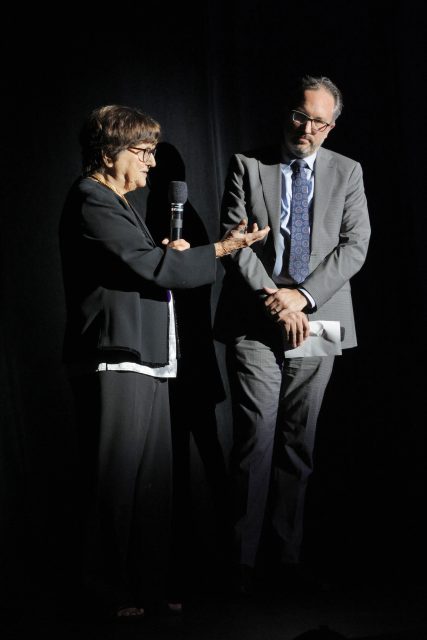 An older woman speaks into a microphone while a man in a suit stands beside her, both illuminated against a dark background.