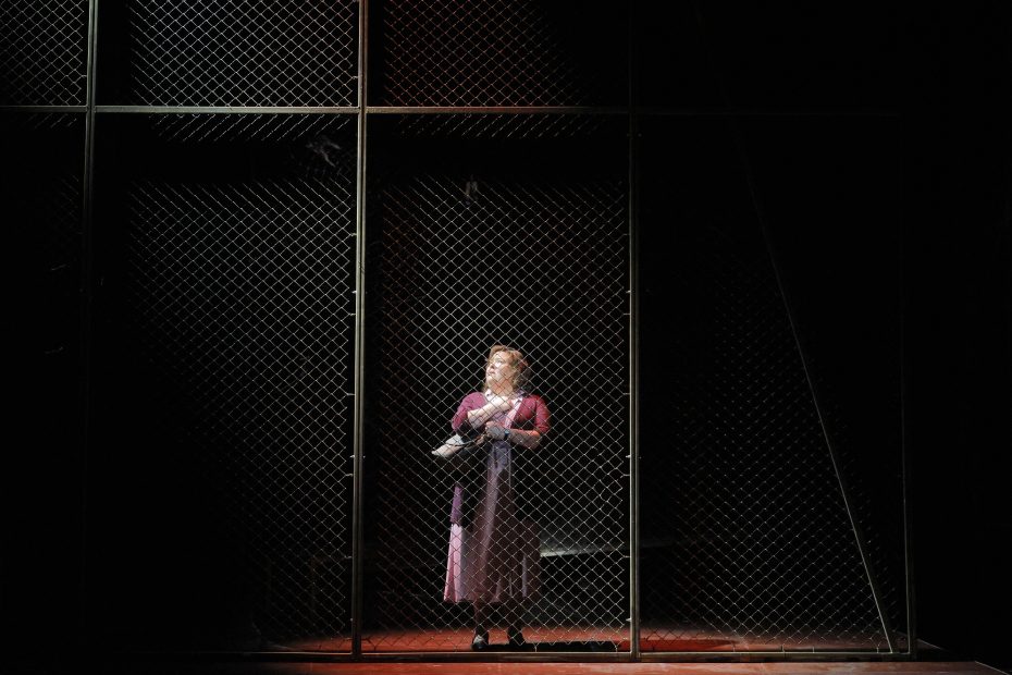 A woman in a long dress stands alone behind a tall chain-link fence on a dimly lit stage, holding a bundle in her arms.
