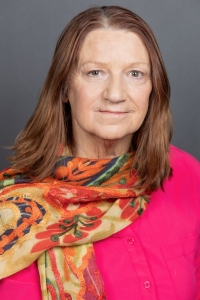 A middle-aged woman with straight brown hair wearing a bright pink top and a colorful patterned scarf, posed in front of a plain gray background.