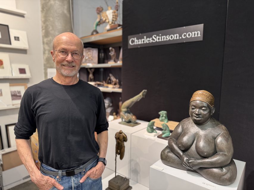 A man stands next to a large seated female sculpture in an art gallery, with other sculptures and a sign reading "CharlesStinson.com" visible in the background.