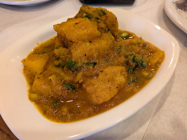 A white dish containing a serving of curried potatoes in a yellow sauce, garnished with herbs, placed on a white table.