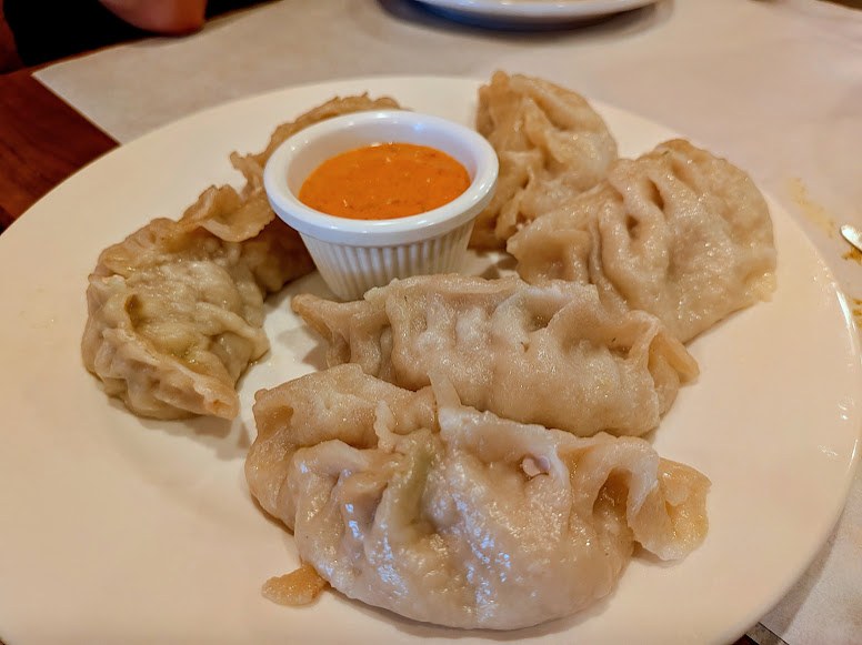 A white plate with five steamed dumplings arranged around a small cup of orange dipping sauce.