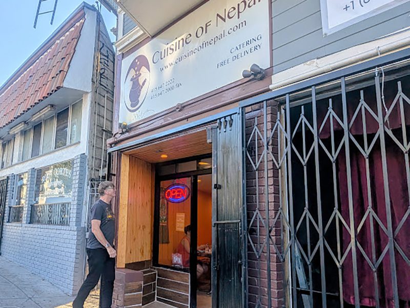 A person stands outside the entrance of Cuisine of Nepal, with an open sign and the business hours clearly visible on the restaurant door.
