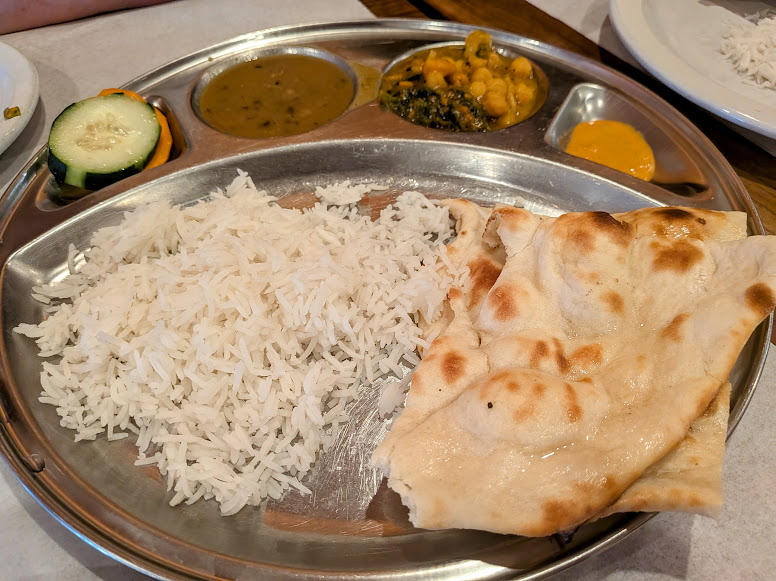 A metal tray with white rice, naan bread, lentil curry, vegetable curry, pickles, and sliced cucumber and carrot.
