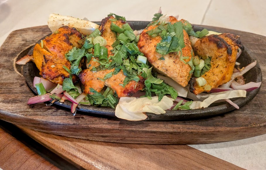 Grilled chicken pieces garnished with chopped cilantro and green onions, served on a bed of sliced onions and cabbage on a wooden platter.