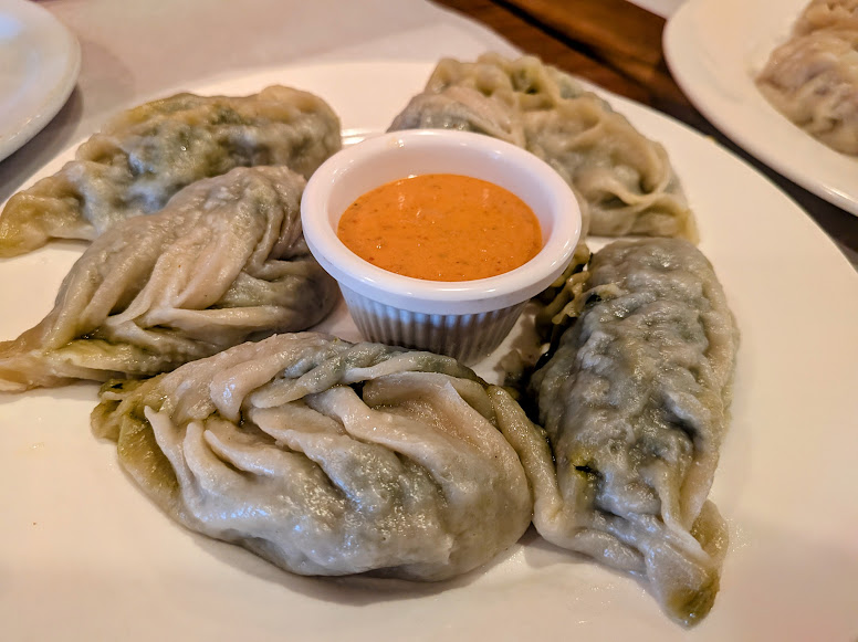 Five steamed dumplings arranged around a small cup of orange dipping sauce on a white plate.