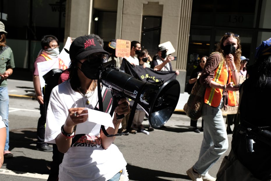 A group of people march on a city street; one person in front speaks into a megaphone while holding a paper, and several others wear masks and safety vests.