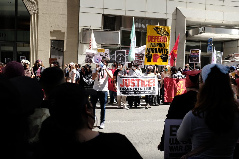 A group of protesters gather on a city street holding signs and banners supporting migrants and justice for Brandon Lee.