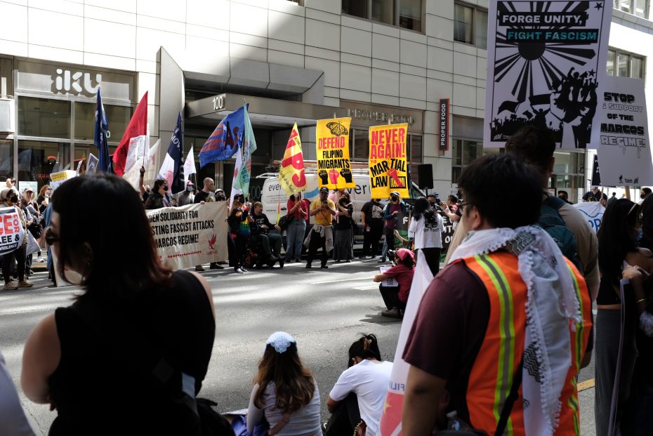 A group of people protest on a city street, holding various signs and banners with messages about unity, migrants' rights, and opposition to fascism and martial law.
