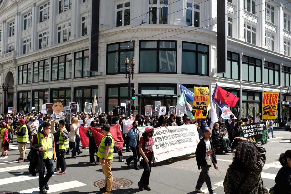 A large group of people march across a city street holding signs and banners during a daytime protest in front of a white multi-story building.