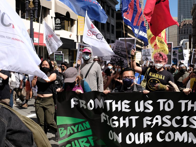 A group of people march in a city street holding a banner reading "From the US to the PH, fight fascist attacks on our communities" and various flags. Many wear face masks.