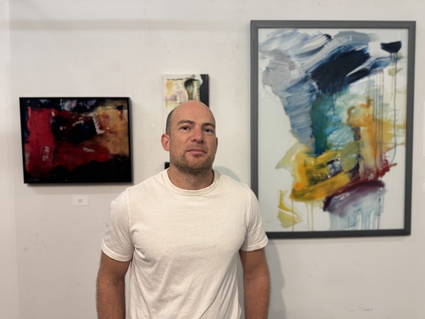 A man in a white t-shirt stands in front of a wall displaying three abstract paintings in an art gallery.