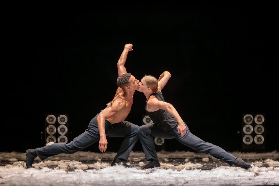 Two dancers in black pants and sleeveless tops perform a dramatic pose on stage, touching faces amid scattered white feathers, with stage lights in the background.