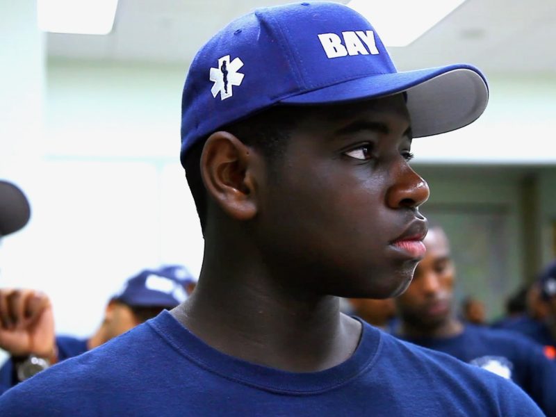 A young man in a blue cap and shirt with "BAY" on the cap looks to the side in a room with others, some appearing to be In the Red team, all dressed in similar attire.