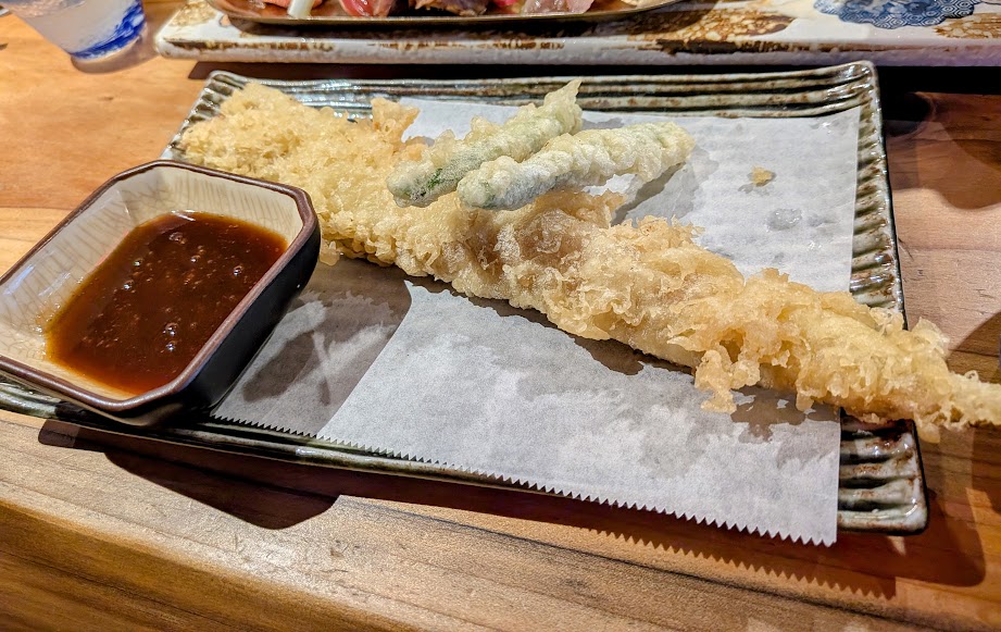 A long piece of tempura and a few smaller tempura vegetables on parchment with a dish of dipping sauce, served on a rectangular plate.