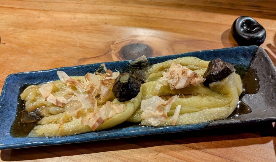 A rectangular blue plate with two pieces of cooked eggplant topped with bonito flakes and sauce, garnished with a dark condiment, on a wooden table.