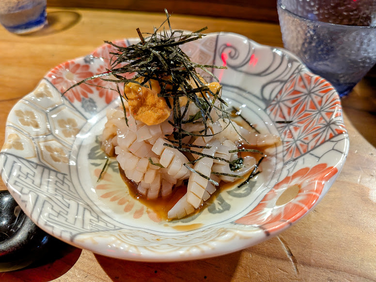 A decorative bowl with sliced squid sashimi topped with sea urchin and shredded nori, served with sauce.
