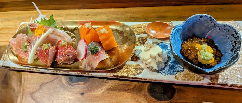 A platter with assorted sashimi, sliced radish, pickled garnish, a dollop of sauce, and a blue bowl of salmon roe topped with wasabi on a rectangular ceramic plate.