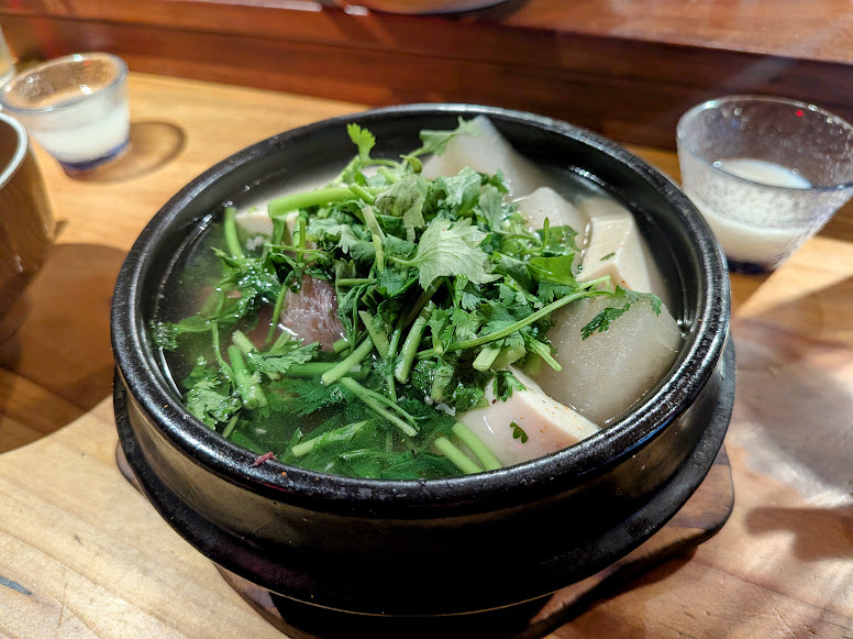 A black bowl of clear soup with herbs, tofu, and vegetables sits on a wooden table, accompanied by a small glass of a milky white beverage in the background.