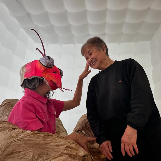 A person in a pink insect costume touches the face of a smiling woman in a black sweater, both inside a white, padded room-like space.