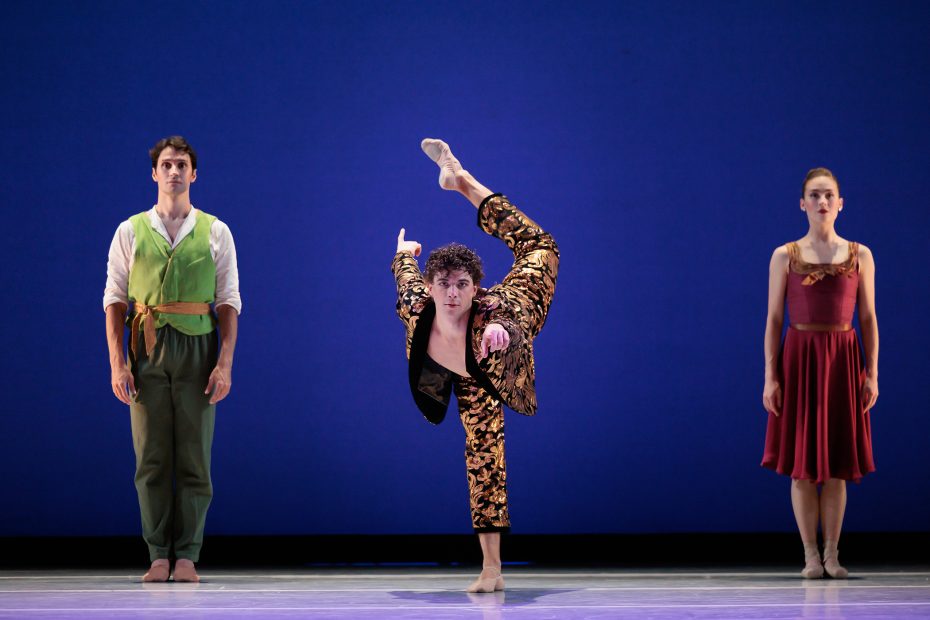Three ballet dancers perform on stage; the center dancer balances on one leg with the other leg extended high, while the two dancers on either side stand upright against a blue backdrop.