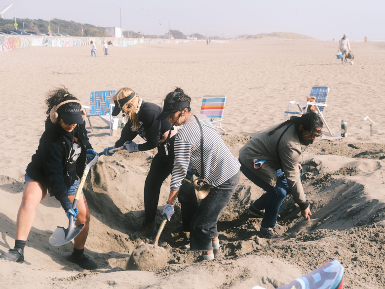 Four people wearing casual clothing dig a large hole in the sand on a beach, with empty chairs and distant figures visible in the background.