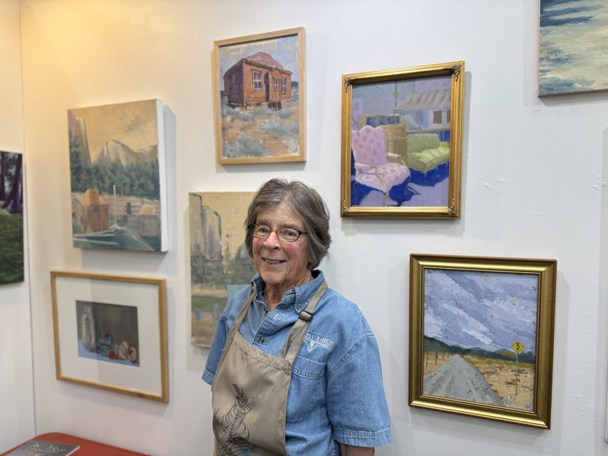 An older woman in glasses and an apron stands in front of a wall displaying several framed landscape and still life paintings.