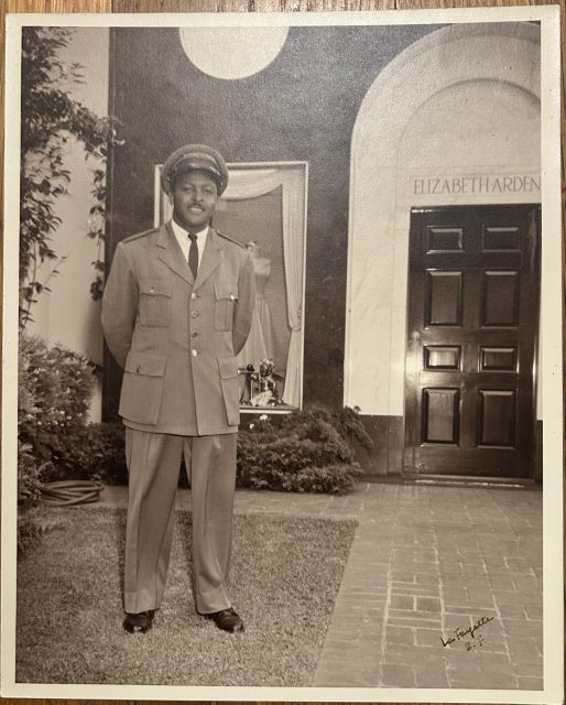 A man in a uniform poses in front of a building with a sign reading "ELIZABETH ARDEN" above a doorway. The photo is black and white and appears to be vintage.