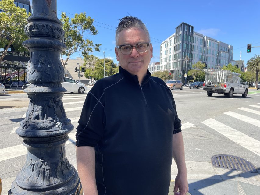 A man wearing glasses and a black pullover stands on a city street corner near a decorative lamppost, with buildings, trees, and cars in the background.