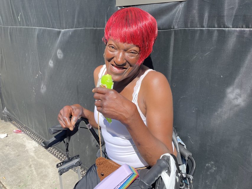 A person with a bright red hairstyle sits in a wheelchair, smiling and holding a green popsicle in one hand, outdoors in sunlight near a black fabric backdrop.