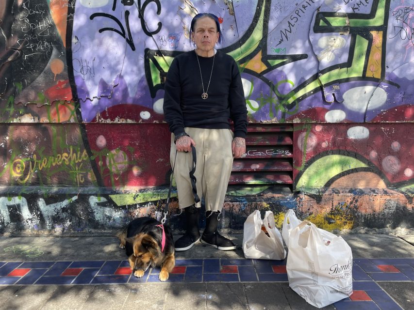 A person sits on a bench in front of a graffiti-covered wall, holding a dog leash with a dog lying beside them and several grocery bags on the ground.