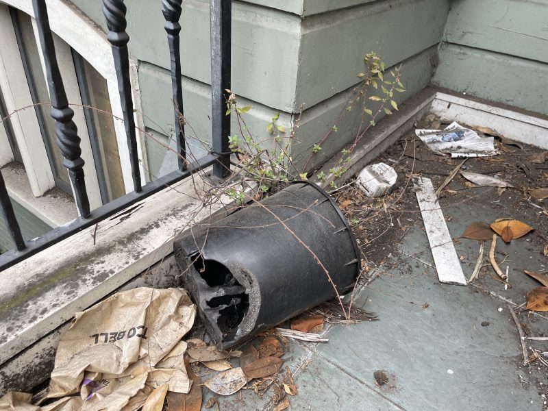 A broken black plastic plant pot lies on its side on a dirty balcony, surrounded by debris, dead leaves, and scraps of paper.