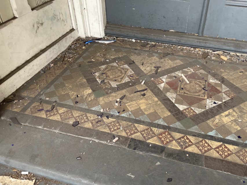 Old, patterned tile floor with debris, dirt, and broken glass scattered near a gray door and weathered white wall.
