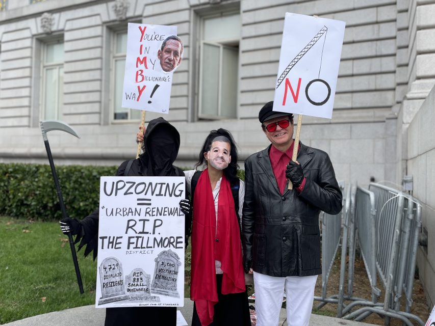 Three people stand outside a building holding protest signs, one person dressed as the Grim Reaper and another wearing a mask, protesting urban renewal and upzoning.