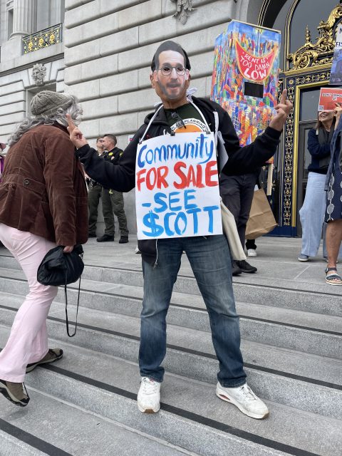 Person wearing a mask and a sign reading "Community for Sale - See Scott" stands on steps outside a stone building among other protesters.
