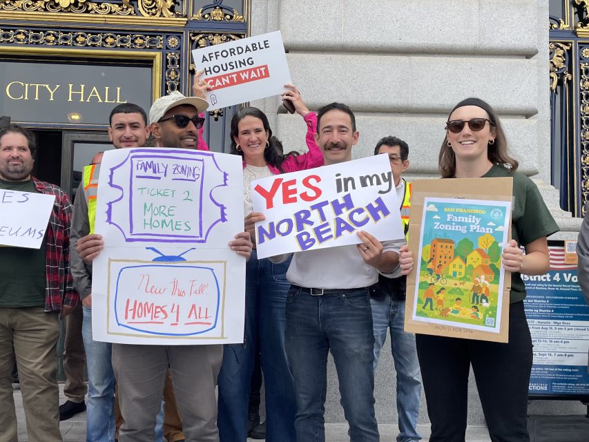 A group of people stand outside City Hall holding signs supporting affordable housing and family zoning plans.