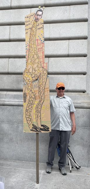 A man in a gray shirt and orange cap stands next to a tall giraffe-shaped protest sign with a person's face and text, in front of a large stone wall.