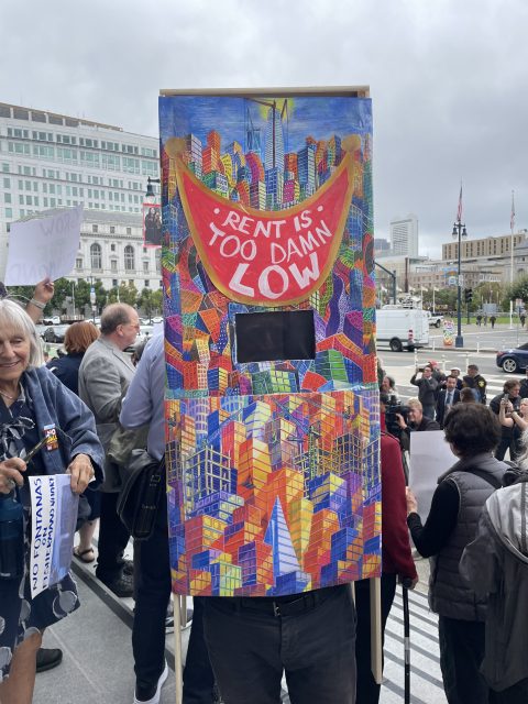 A person stands at a protest holding a colorful sign resembling a cityscape with the words "RENT IS TOO DAMN LOW" in large letters. People and buildings are visible in the background.