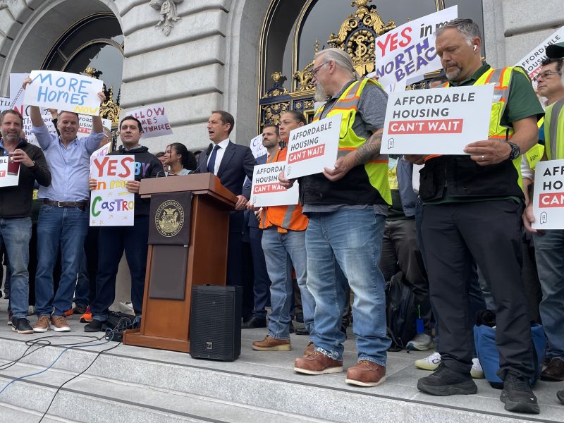 A group of people stand on city hall steps holding signs in support of affordable housing and more homes, with a podium and speaker in the center.