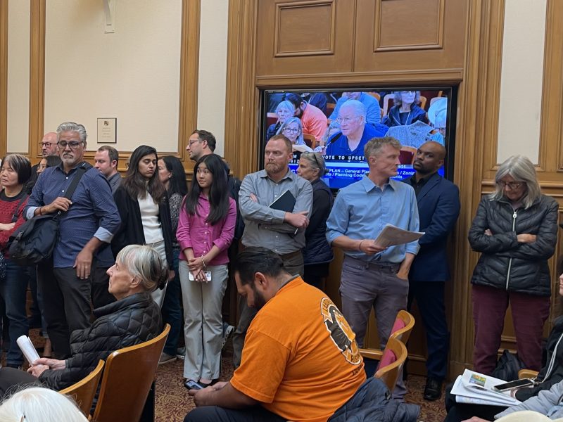 A group of people stand and sit in a meeting room; a screen in the background displays a man speaking at a podium.