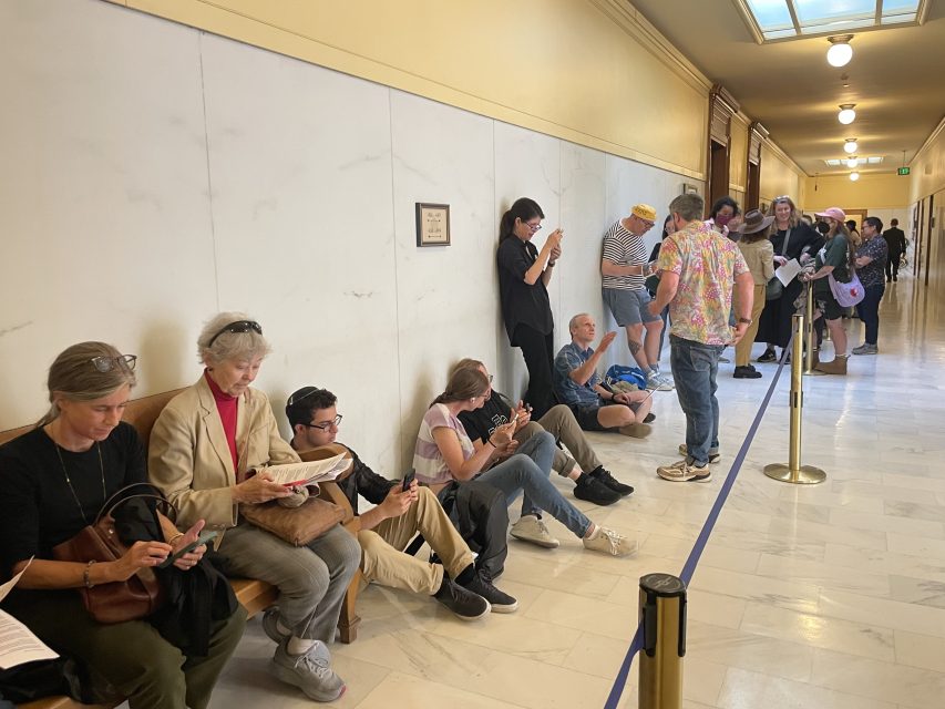 People sit and stand in a hallway, some using their phones or reading, while waiting in a line near a wall with a rope divider and a marble floor.