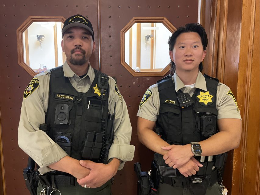 Two uniformed officers wearing body cameras and name badges stand side by side in front of double wooden doors with hexagonal windows.