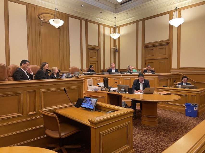 A group of people sit at desks and benches in a wood-paneled courtroom, some with laptops and papers, during a formal proceeding.