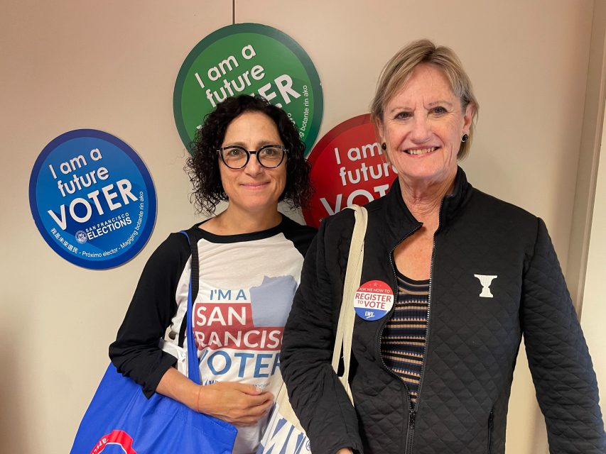 Two women stand in front of a wall with "I am a future voter" signs. One wears glasses and a "San Francisco voter" shirt, while the other—sporting a black jacket and "Register to Vote" sticker—shows support for the upcoming recall election.