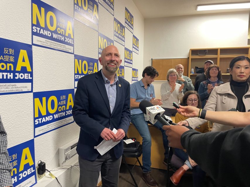 A man in a suit stands smiling at a recall press event, surrounded by people and "No on A, Stand with Joel" signs covering the wall behind him as reporters hold microphones toward him.