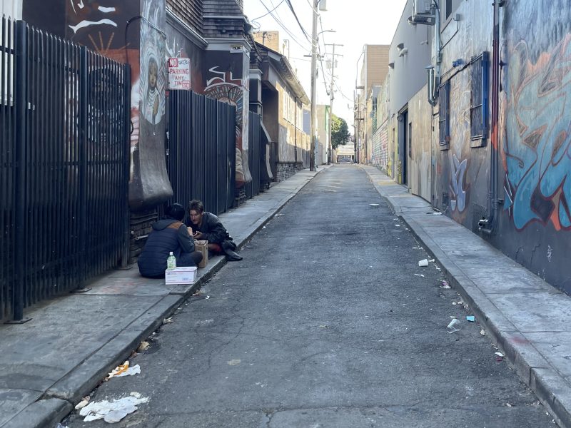 Two people sit on the sidewalk in an urban alley lined with graffiti-covered walls and scattered trash.