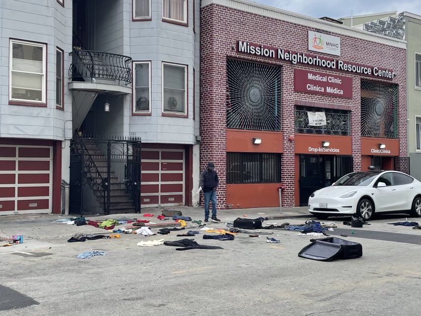 A person stands in front of a building with scattered clothing and suitcases on the ground near parked cars outside the Mission Neighborhood Resource Center.